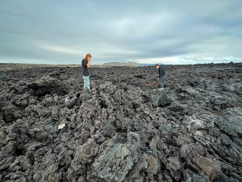 Two individuals walk across a rugged lava field under a cloudy sky. A small white stone is visible among the dark rocks.