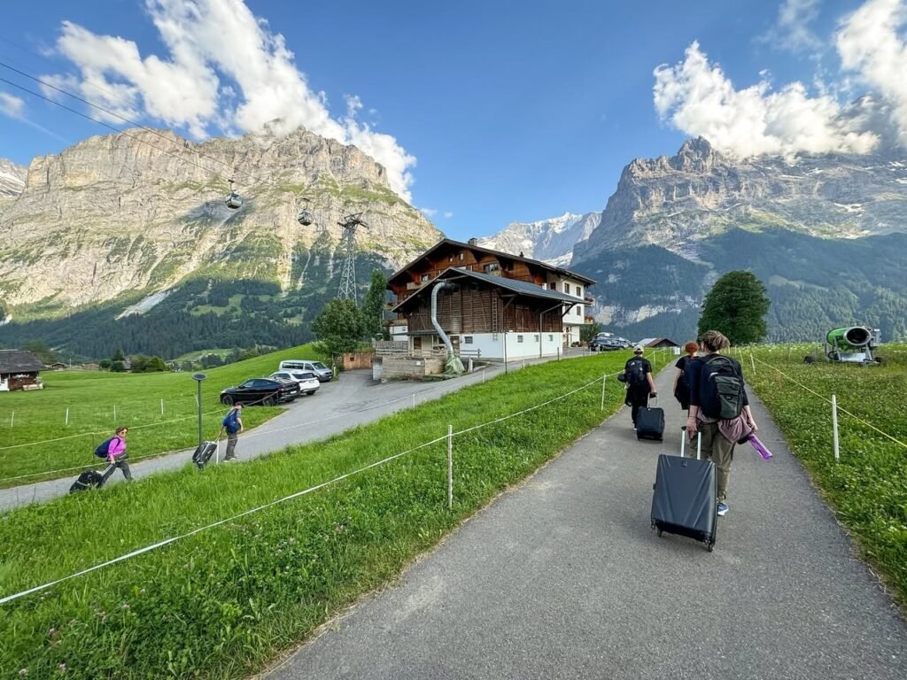 Travelers with luggage walk on a paved path through a green alpine meadow toward a traditional wooden chalet, set against a backdrop of towering rocky mountains and a bright blue sky.