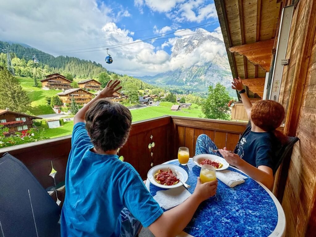 A family enjoys breakfast on a wooden balcony overlooking green mountain valleys and a cable car. They watch the scenery with juice and fruit bowls on a blue table.