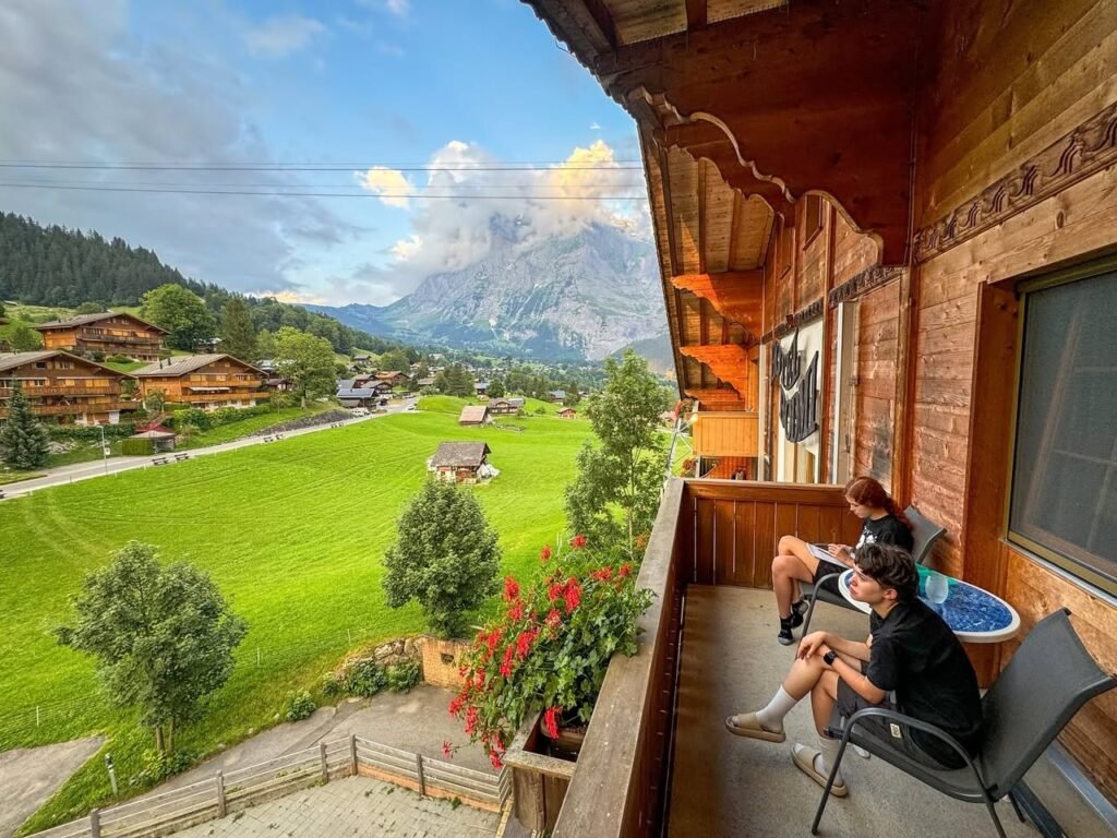 View of a small village and green valley from the stone porch of a mountain lodge. People relax on the wooden balcony with red flowers.