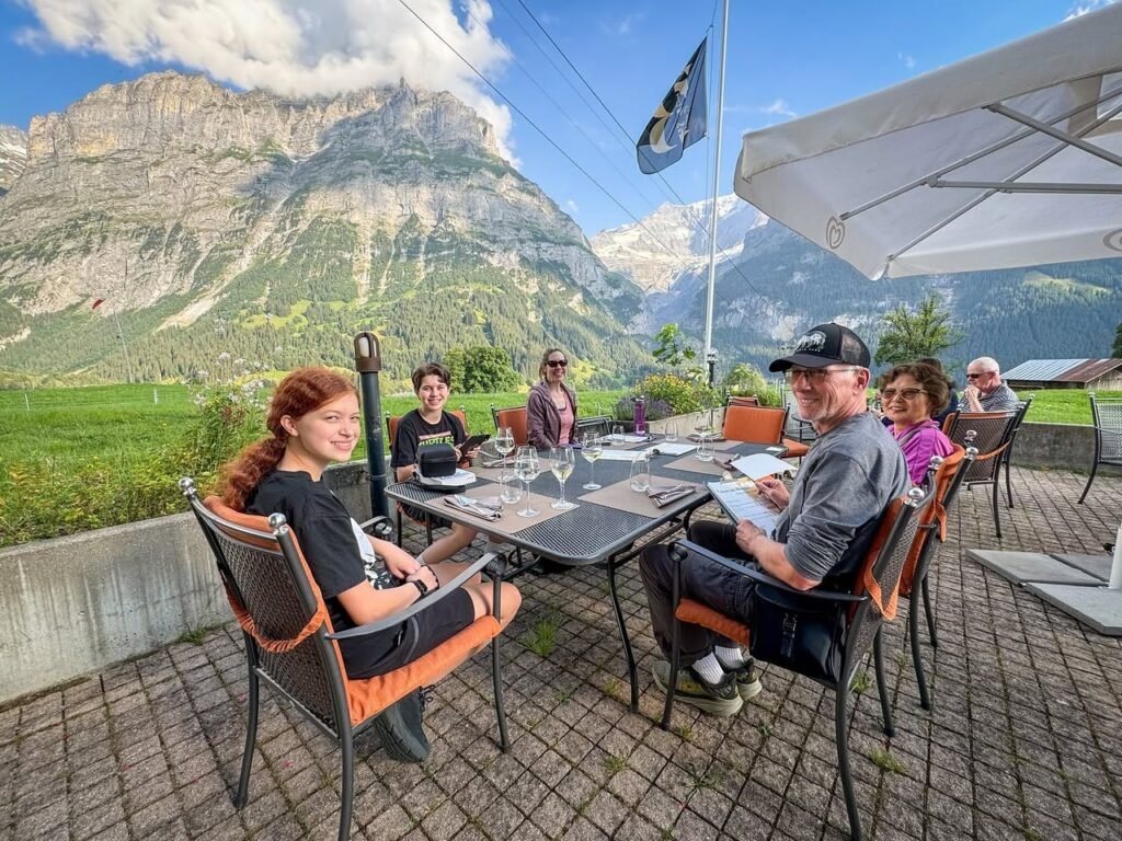 A gathering of travelers on a scenic alpine terrace enjoys a traditional meal against a backdrop of majestic mountains. The event coincides with May 1st.