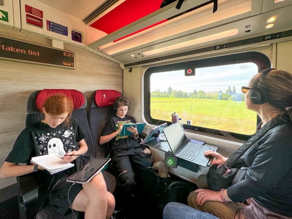 A family enjoys reading and working with tablets on a train. The interior features red seats and a view of green fields outside.