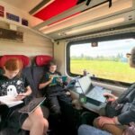 A family enjoys reading and working with tablets on a train. The interior features red seats and a view of green fields outside.