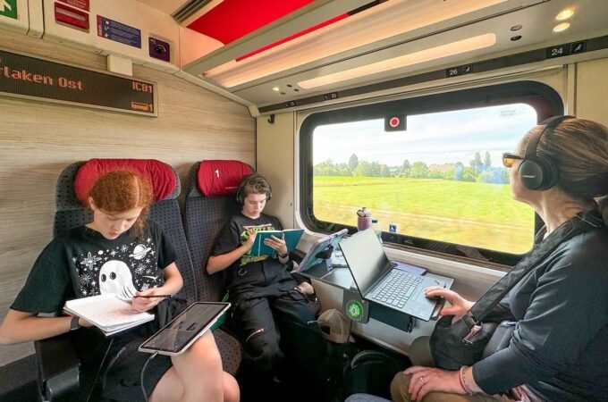 A family enjoys reading and working with tablets on a train. The interior features red seats and a view of green fields outside.