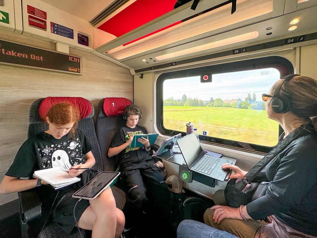 Hotel Bodmi A family enjoys reading and working with tablets on a train. The interior features red seats and a view of green fields outside.