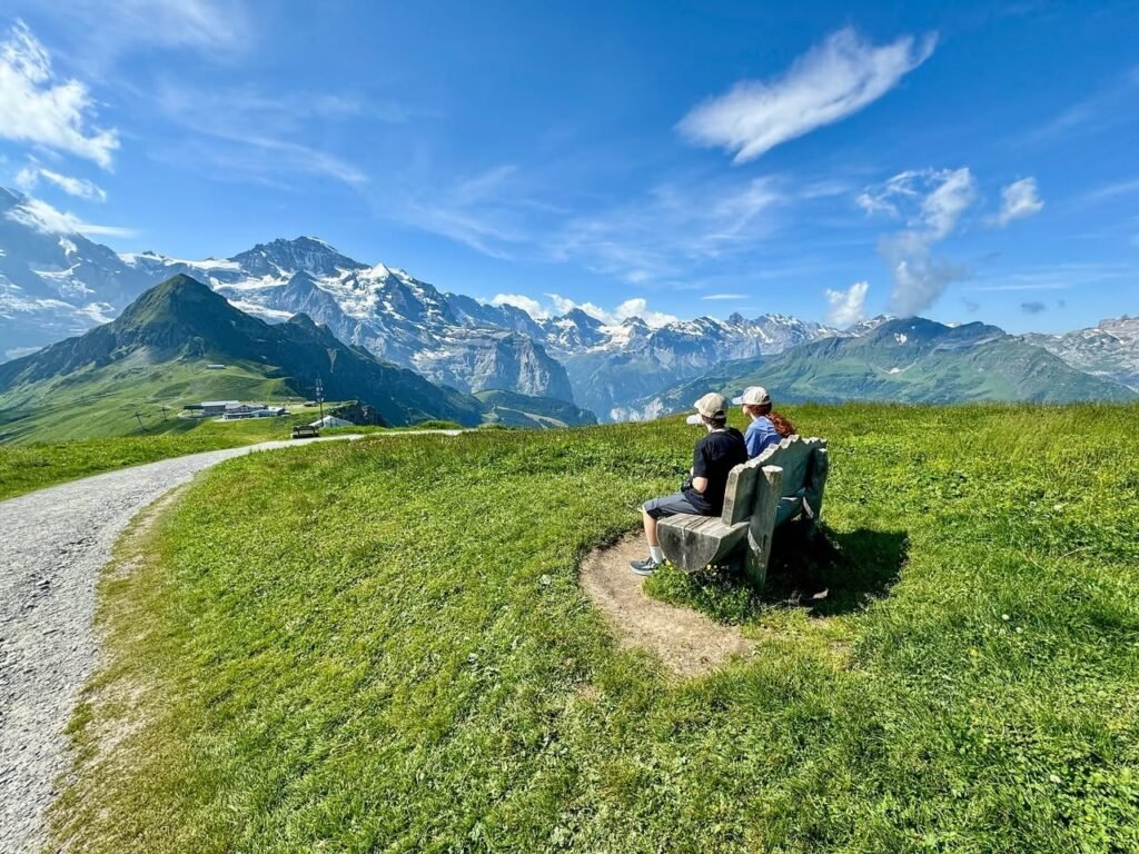 Two people sit on a wooden bench overlooking a grassy field and snowy mountain range under a blue sky with scattered clouds.