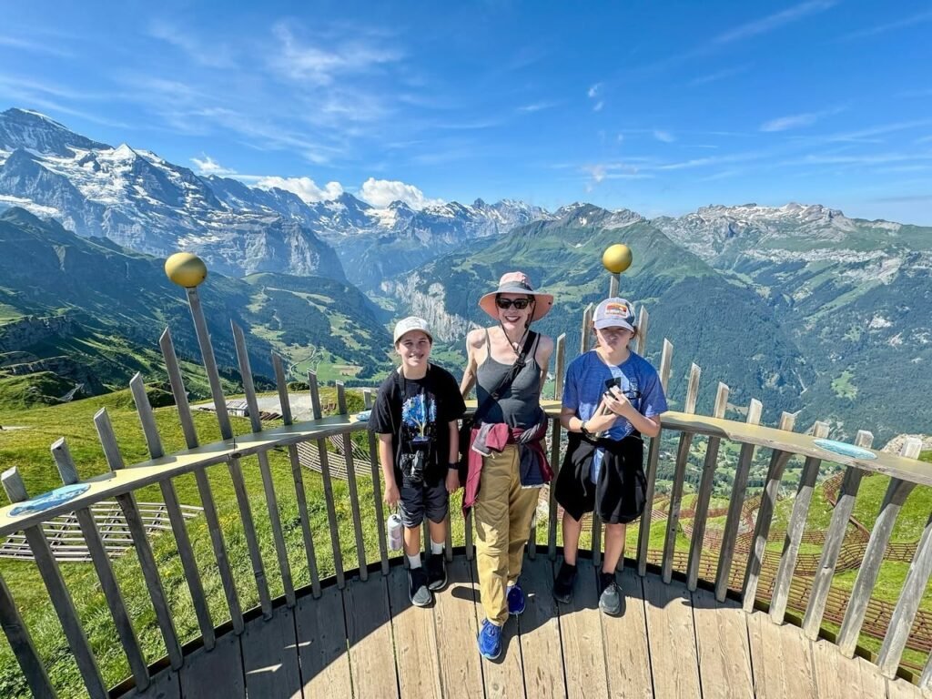 A group of travelers stands on a wooden mountain deck surrounded by a stunning alpine landscape. Snow-capped peaks and a bright blue sky complete the scenic view.