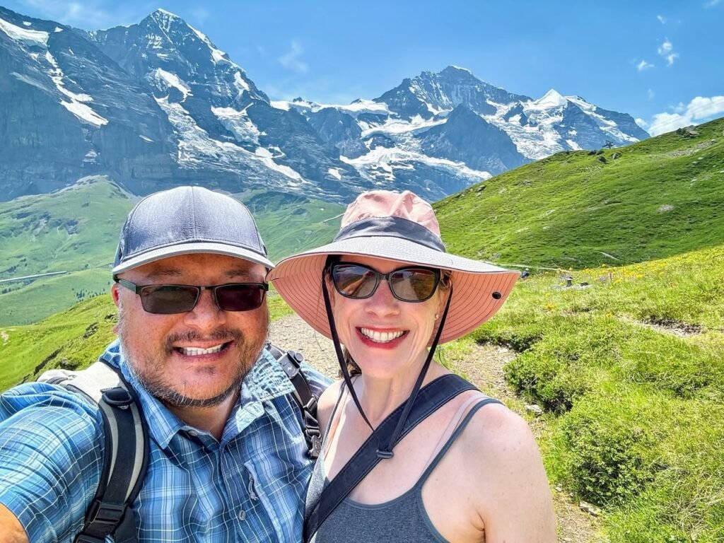 A couple taking a selfie while hiking in the mountains with snow-capped peaks in the background. The second image shows a close-up of one hiker with a mountain view behind, and the third displays lush green alpine terrain.