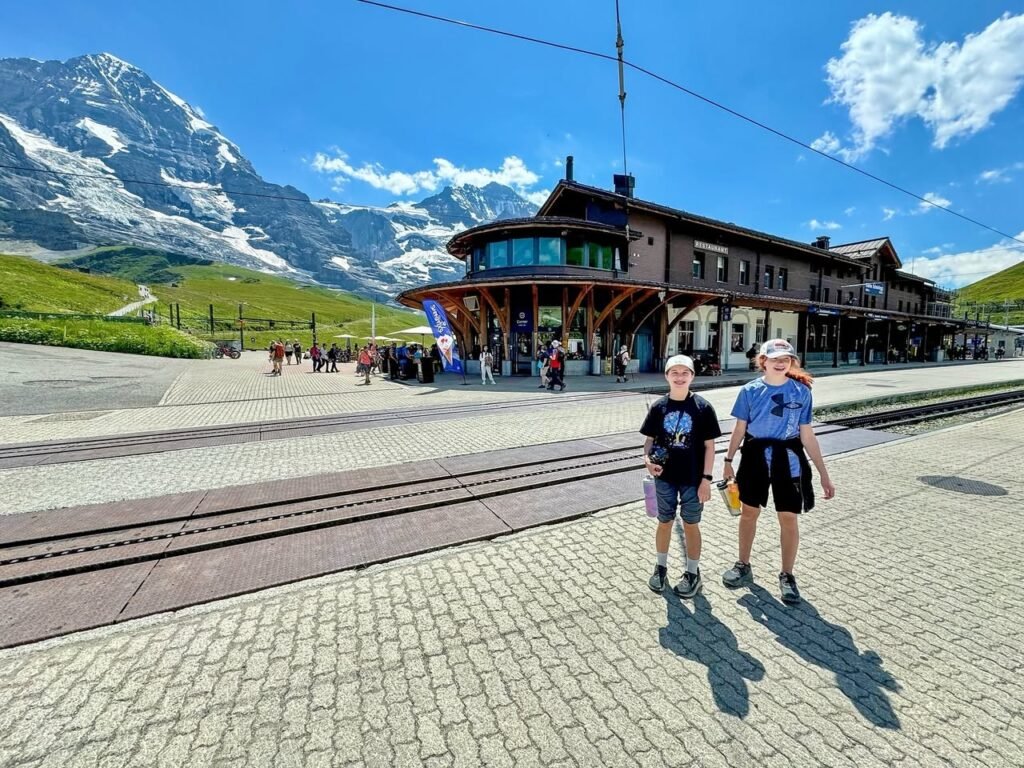 Two visitors walk along the paved platform of the Grindelwald Jag Shuttle station, with snow-capped mountains rising in the background.