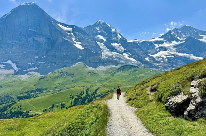 A hiker walks a gravel path through a green alpine meadow. Snow-capped mountains and a blue sky form the background.
