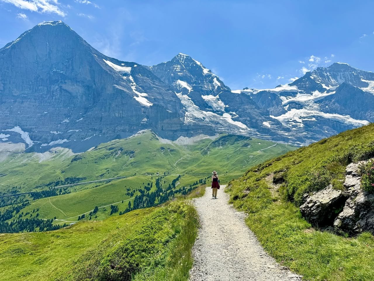 A hiker walks a gravel path through a green alpine meadow. Snow-capped mountains and a blue sky form the background.