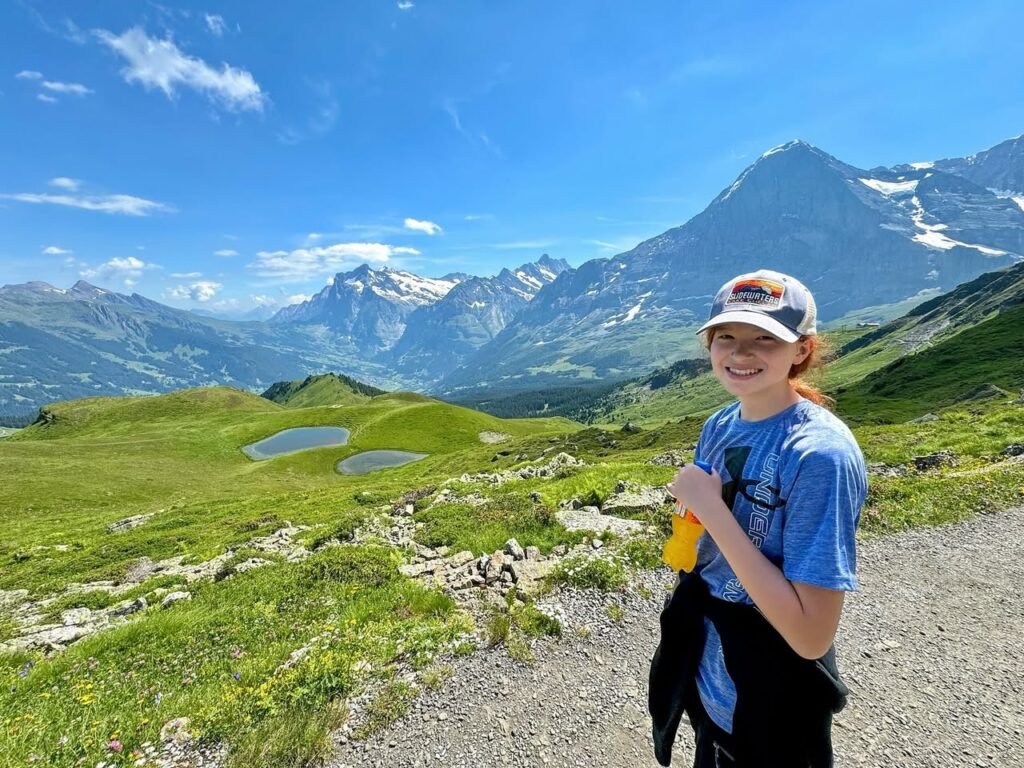 A hiker stands on a gravel path in a lush green alpine valley with snow-capped mountains in the background.
