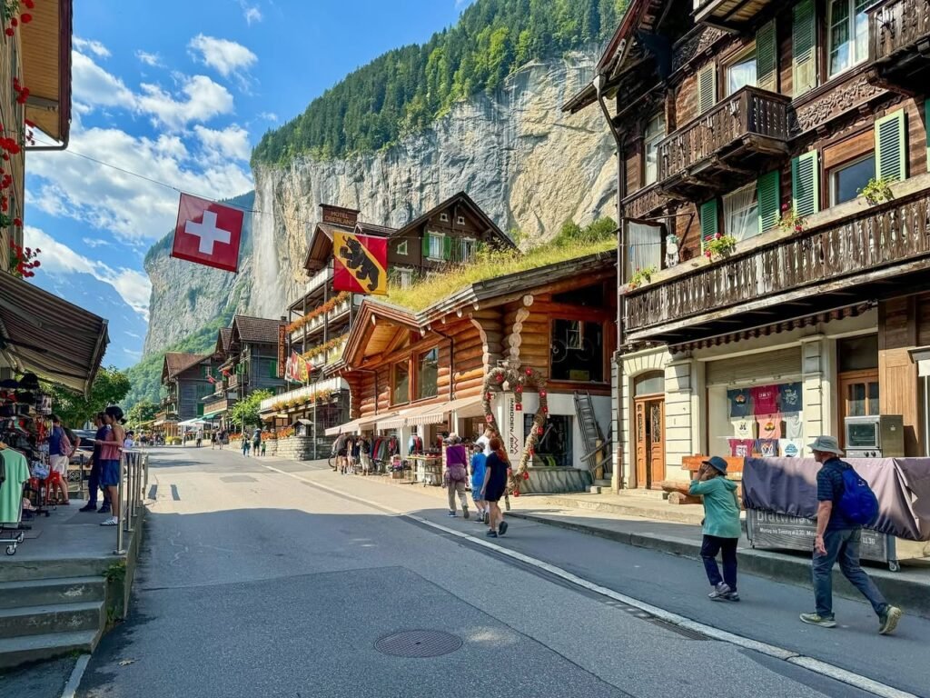 Traditional wooden houses with green shutters and Swiss flags in a picturesque mountain village. A narrow street with people walking under a bright, clear sky.