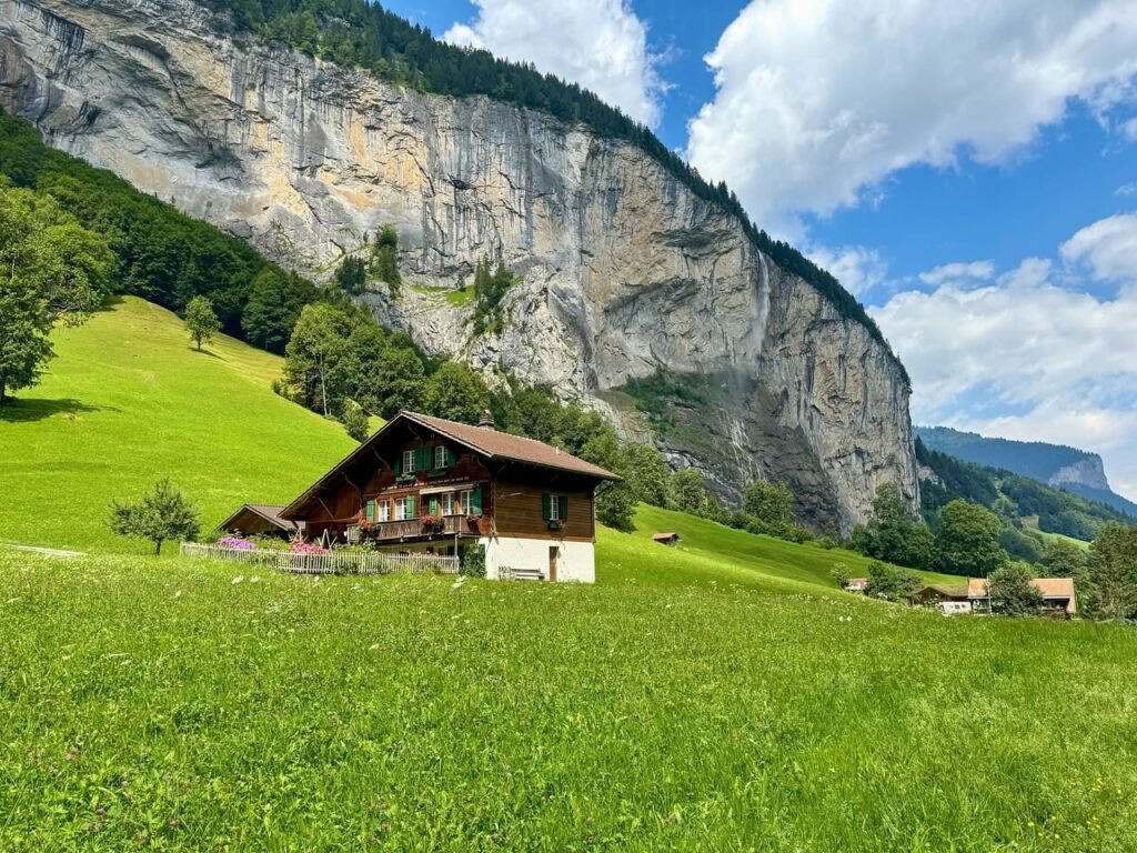 A Swiss-style farmhouse sits in a lush green valley beneath towering limestone cliffs and a blue sky.