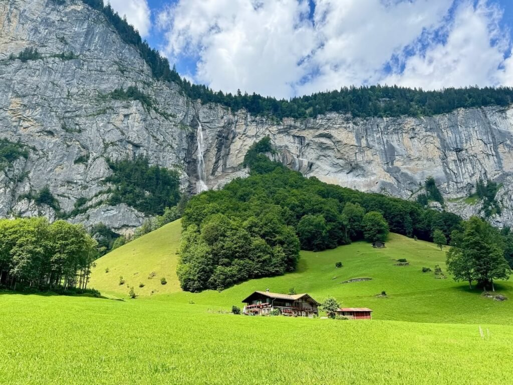 A rustic barn sits alone in a bright green meadow beneath towering rocky cliffs and a partly cloudy sky.