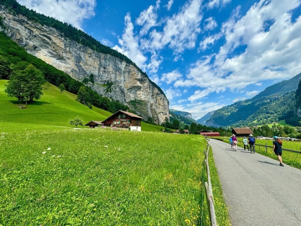 A paved walking path winds through vibrant green alpine meadows along a rocky cliffside. The bright blue sky features scattered white clouds above the valley.