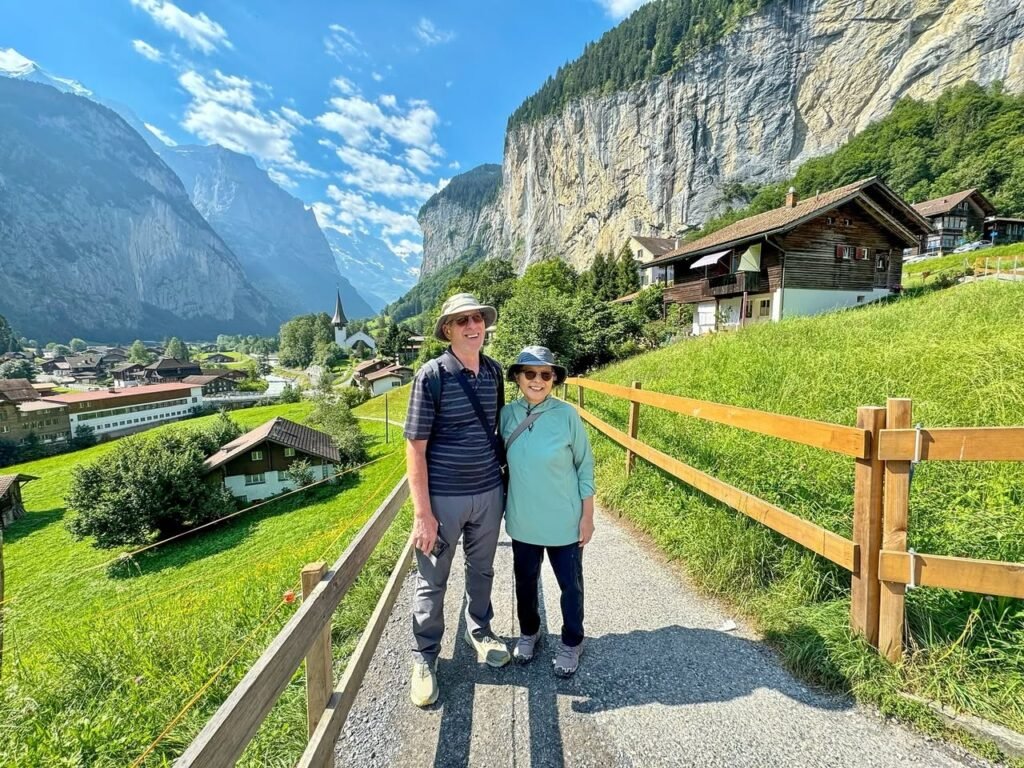 Two people walking on a gravel path beside a wooden fence with a mountain view in the background.