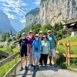 A group of hikers poses on a scenic mountain path surrounded by lush greenery and towering cliffs. Traditional alpine houses and a distant church spire complete the picturesque landscape.
