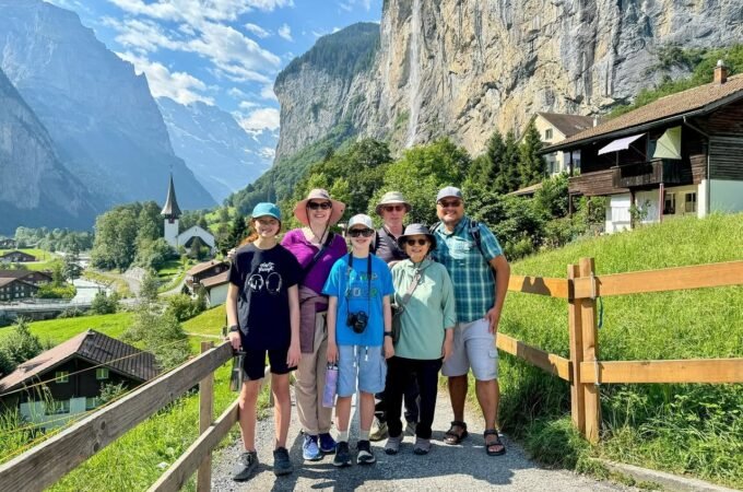A group of hikers poses on a scenic mountain path surrounded by lush greenery and towering cliffs. Traditional alpine houses and a distant church spire complete the picturesque landscape.