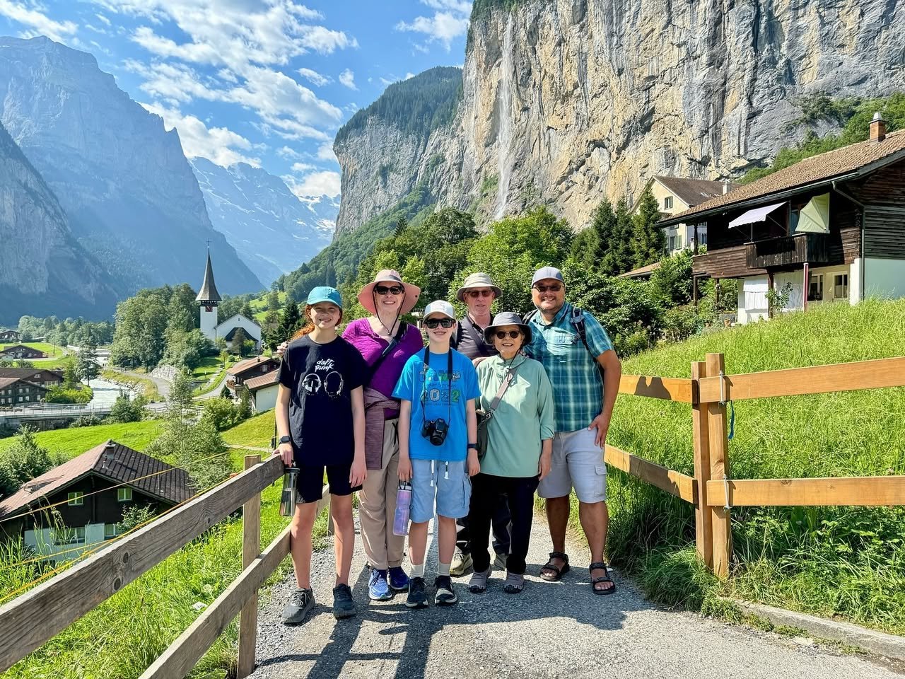 Lauterbrunnen AKA Rivendell A group of hikers poses on a scenic mountain path surrounded by lush greenery and towering cliffs. Traditional alpine houses and a distant church spire complete the picturesque landscape.