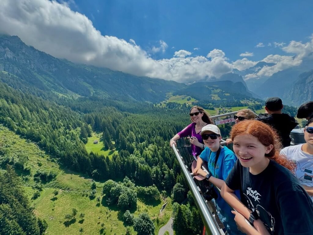 People enjoying a scenic overlook with lush green mountains and forests. A vibrant blue sky with fluffy white clouds caps the landscape.