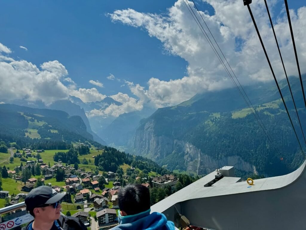 Passengers view panoramic mountain scenery and a valley village from a gondola, framed by cable lines against a partly cloudy blue sky.