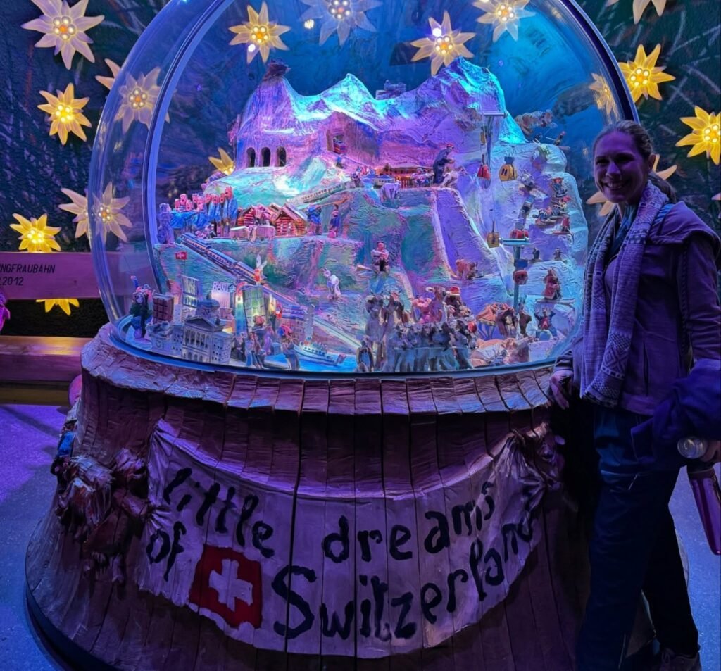 A person standing in front of a large illuminated snow globe titled Little Dream of Switzerland. The glass dome contains a colorful miniature model with stars around it.