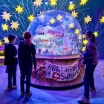 Karaoke Gondola Visitors observe a large illuminated snow globe labeled 'A Little Dream of Switzerland' surrounded by glowing yellow stars in a darkened exhibit space.