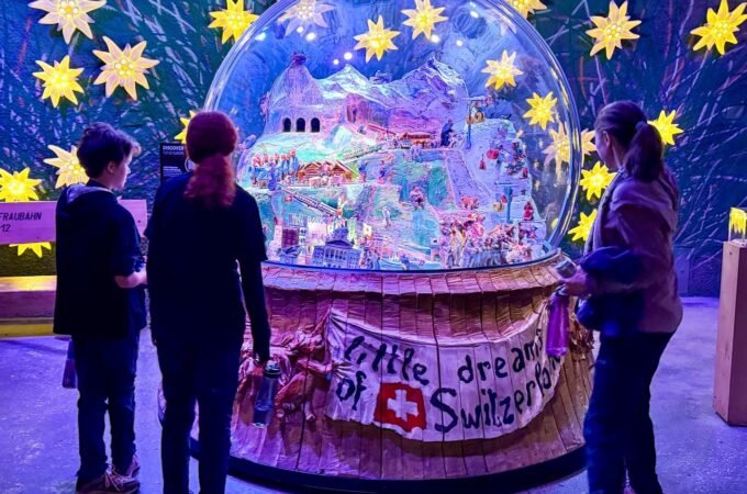 Visitors observe a large illuminated snow globe labeled 'A Little Dream of Switzerland' surrounded by glowing yellow stars in a darkened exhibit space.