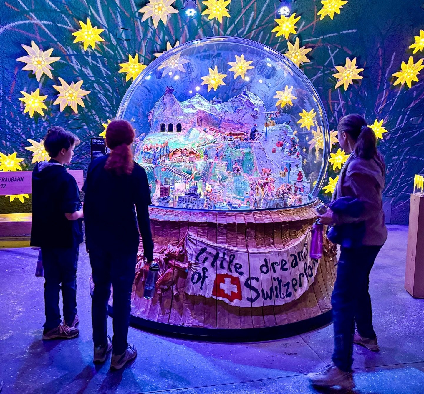 Alpine Sensation Visitors observe a large illuminated snow globe labeled 'A Little Dream of Switzerland' surrounded by glowing yellow stars in a darkened exhibit space.