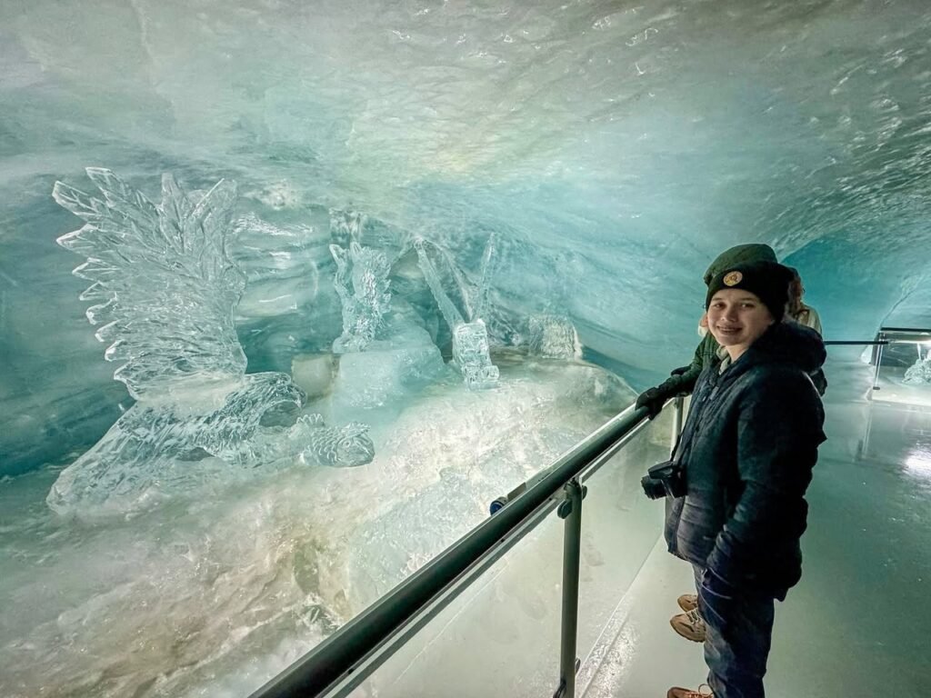 A person stands inside a large ice sculpture chamber, observing detailed carvings and translucent blue ice walls.