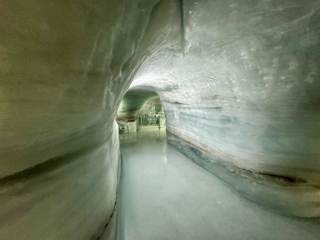 View looking down a narrow ice tunnel with a calm water channel. The smooth surfaces suggest a frozen or icy environment.