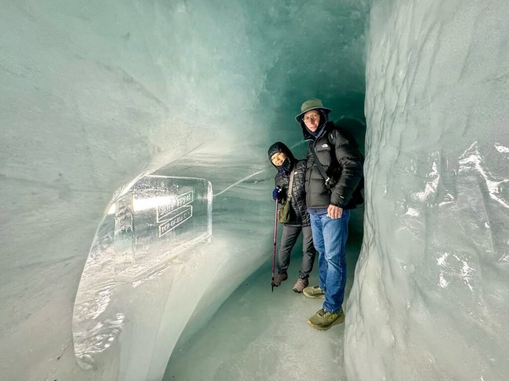 Two individuals explore a vibrant blue ice tunnel carved through a glacier, highlighting the surreal beauty of frozen water.
