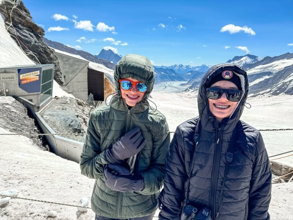 Two travelers in winter gear smiling on a paved view platform at the end of a glacier. Snow-covered mountains and a steel visitor container are visible under a blue sky.