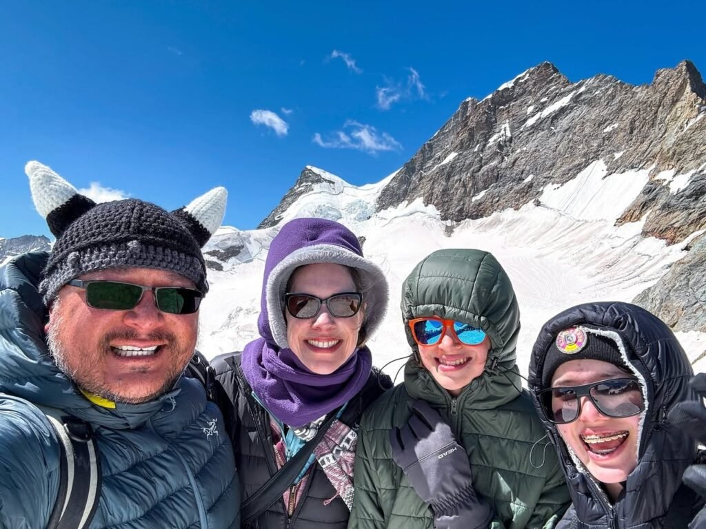 A group of four hikers posing for a smiling selfie on a snow-covered mountain. Wearing winter gear and sunglasses under a bright blue sky.