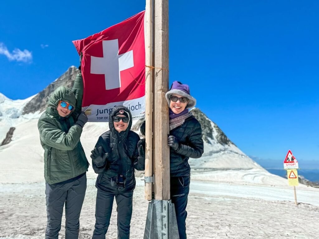 See a group proudly holding a Swiss flag at a high-altitude mountain peak. The background features snowy alpine terrain under a bright blue sky.