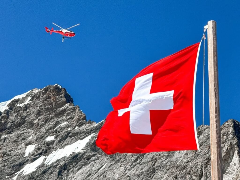 A close-up of a red and white Swiss flag waving on a wooden pole against a backdrop of rocky mountain peaks and clear blue sky.