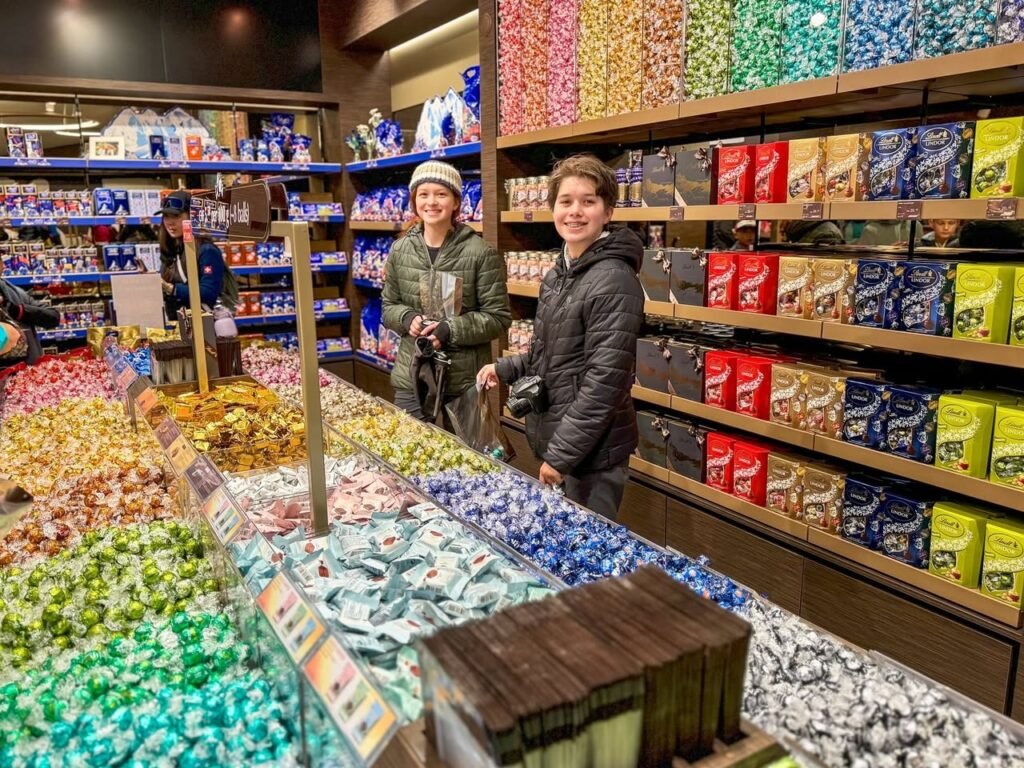 Two people smiling in a candy store surrounded by shelves of colorful chocolate bars and wrapped candies.