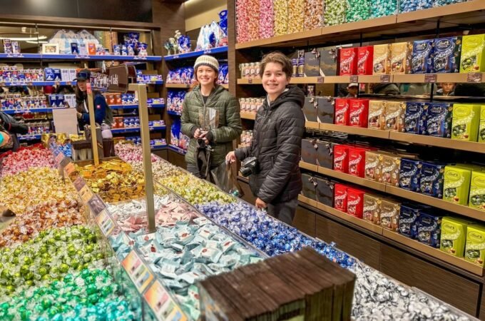 Two people smiling in a candy store surrounded by shelves of colorful chocolate bars and wrapped candies.