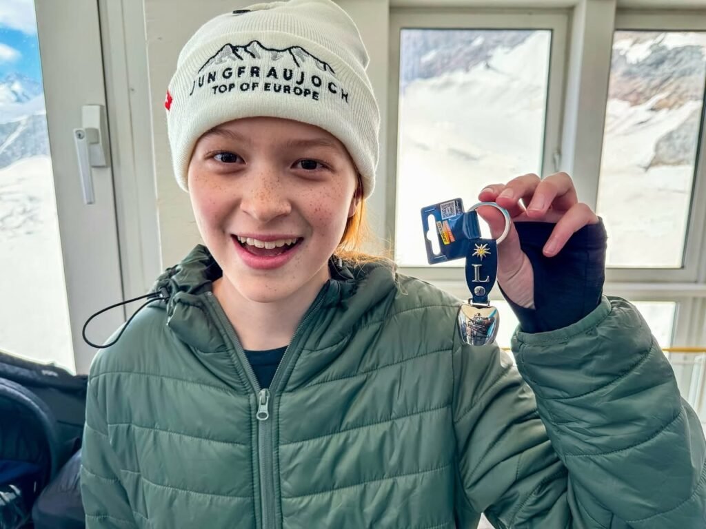 A smiling person wearing a Jungfraujoch beanie and puffy jacket inside a train, displaying a small blue souvenir. Snow-covered mountains are visible through the window.