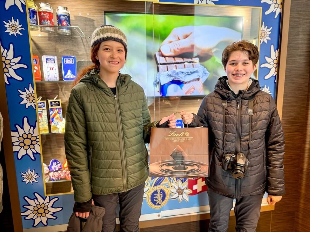Two people in winter jackets hold a branded shopping bag in front of a chalet-style gift shop display.