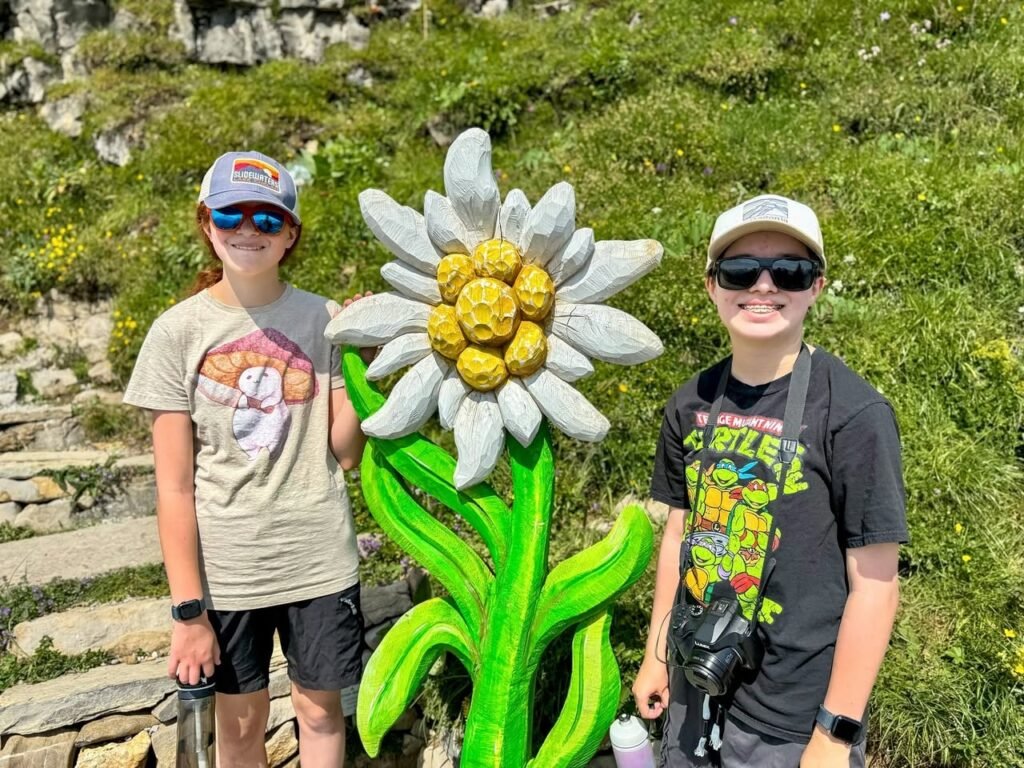 Three individuals wearing caps and sunglasses stand next to a large, colorful flower sculpture on a mountain trail.