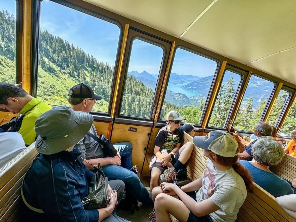Passengers enjoy a panoramic view of dense forest and blue mountains from the windows of a wooden train car.