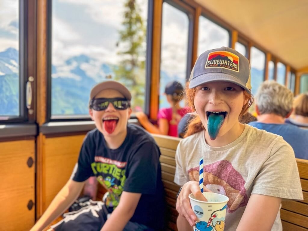Children playfully licking blue ice cream on a train with mountain views outside.