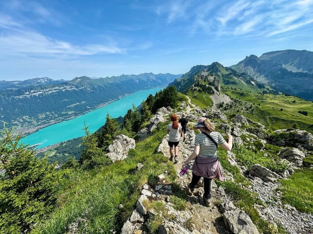 Two trail walkers hiking a rocky ridge as a stunning turquoise lake stretches below, offering sweeping mountain views on a bright, sunny day.