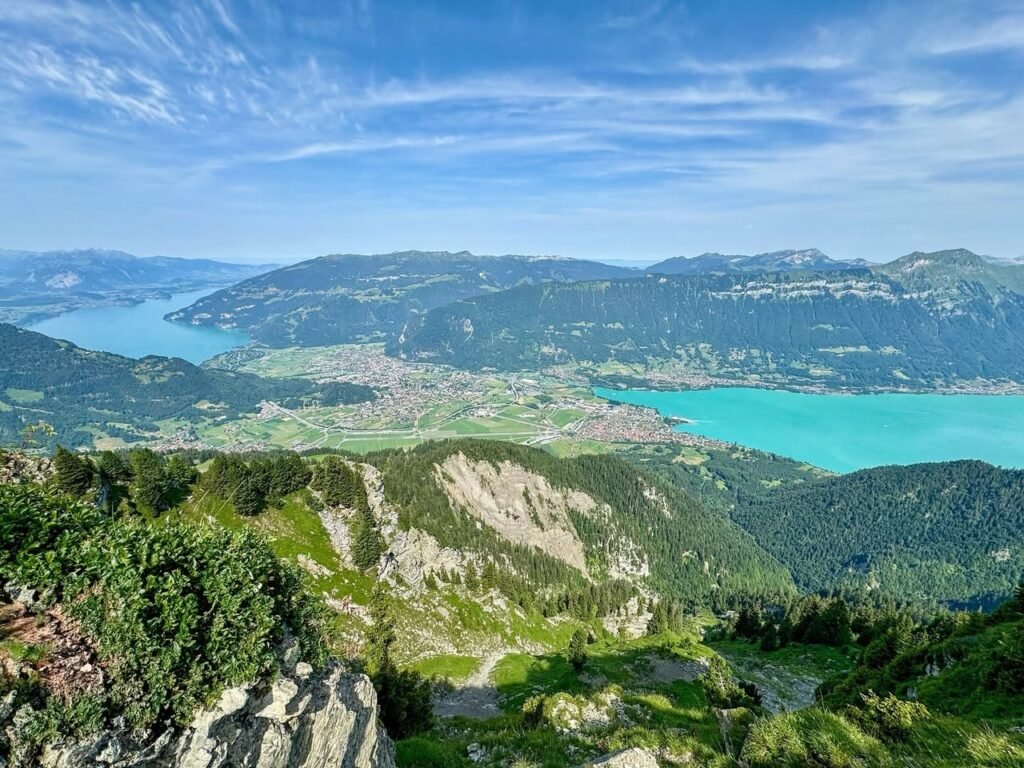 A panoramic view of a green alpine valley featuring a bright turquoise lake and distant mountains under a blue, streaky sky.
