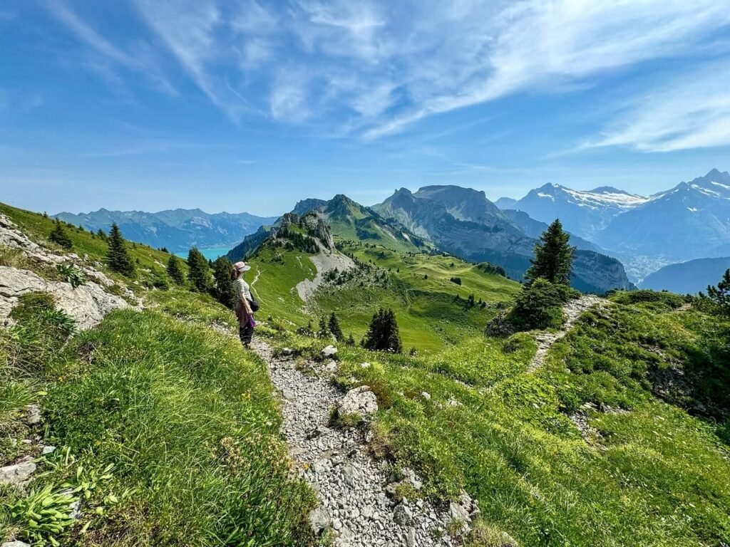 A trail winds through lush green alpine meadows with scenic mountain and lake views under a blue sky.
