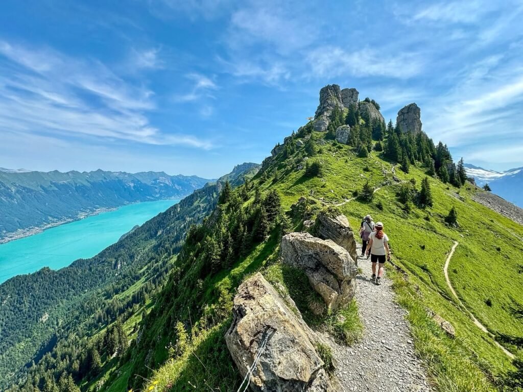 Hikers walk along a rocky mountain trail with a scenic view of a turquoise lake.