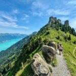 Pfingstegg Hikers walk along a rocky mountain trail with a scenic view of a turquoise lake.