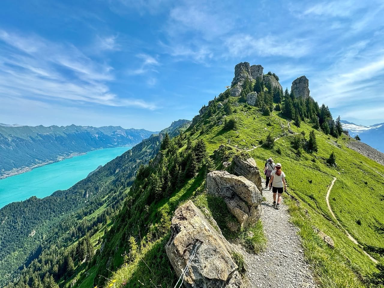 Schynige Platte Hikers walk along a rocky mountain trail with a scenic view of a turquoise lake.